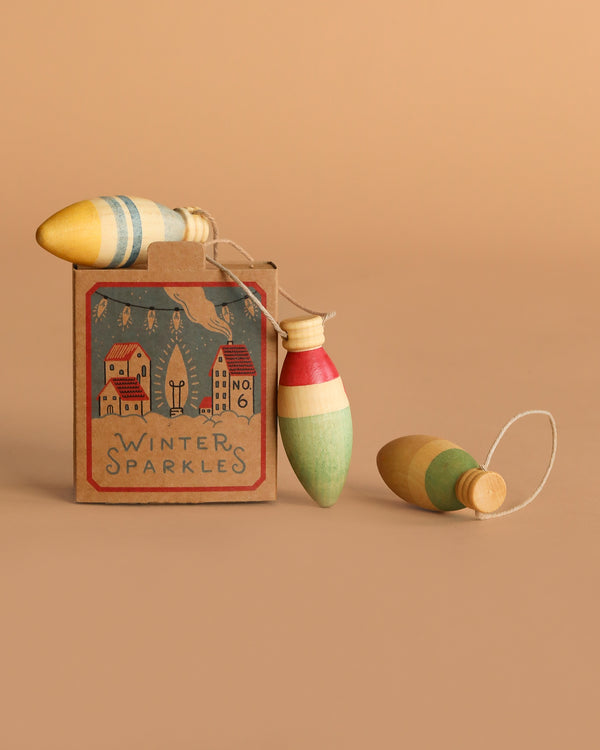 The Grapat Ornament Set - Winter Sparkles features three vintage-style spinning tops with colorful stripes, packaged in a festive box decorated with winter scenes—perfect as hand-painted ornaments or cheerful holiday decorations.