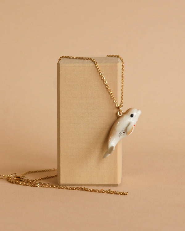 A White Beluga Necklace with a handcrafted porcelain dolphin pendant is displayed hanging over a rectangular beige box. The background is a light beige color, creating a minimalist and oceanic elegance in the presentation.