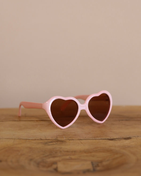 The Minikane Doll Accessories Heart Sunglasses, with pink frames and dark lenses, elegantly rest on a wooden surface, inspired by chic French design against a neutral backdrop.