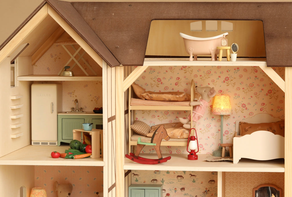 The Maileg Fully Furnished Farmhouse Set + Surprise Gift includes a wooden dollhouse complete with animal figurines, featuring a kitchen, a bedroom with bunk beds, a rocking horse, a lamp, and a bathroom with a clawfoot tub. Each room is adorned with floral wallpaper and petite furniture, enhanced by adorable Maileg Accessories and delightful Maileg Mouse Furniture.