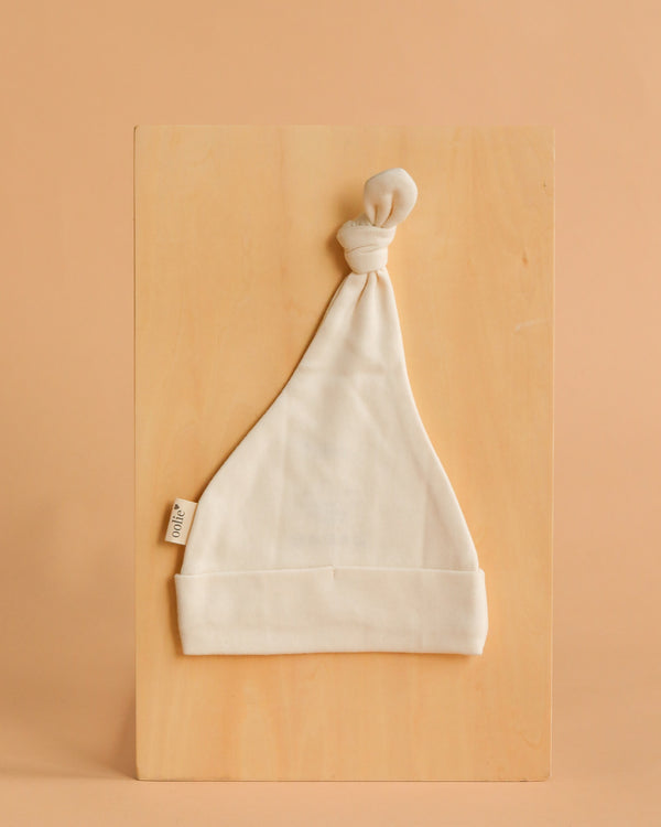 The Organic Baby Hat – Natural, a white GOTS-certified top knot hat with a small fabric tag on the left, is laid flat on a light wooden board against a beige background—ideal for fans of newborn organic clothing.