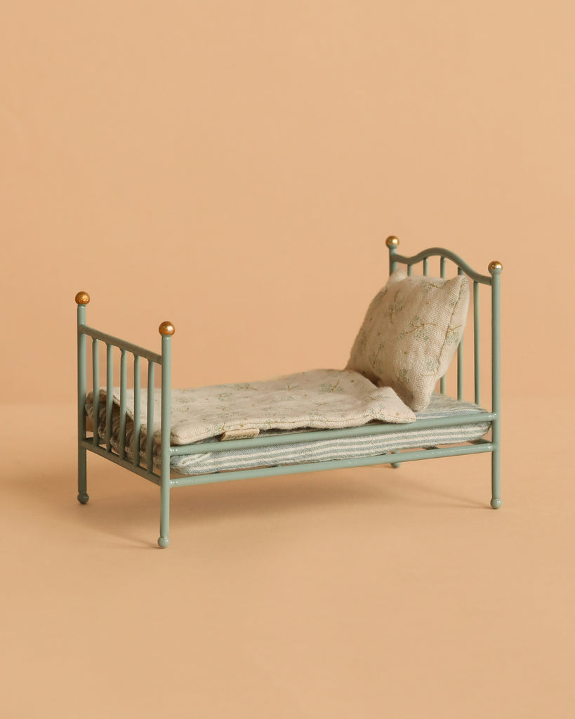 The Maileg Vintage Single Bed, Mouse - Mint features a pale green frame with gold-tipped posts, a floral mattress, and matching pillow—perfect for Maileg mice. Ships in one week. Pictured against a beige background.