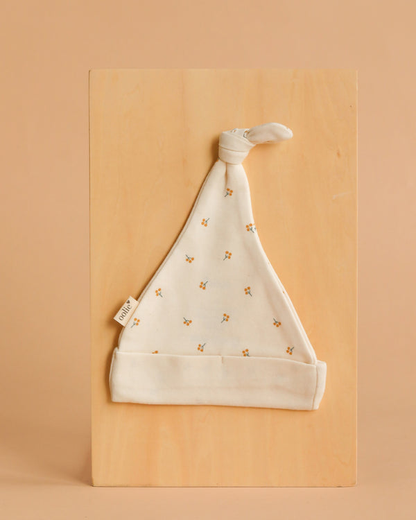 The Organic Baby Hat – Gold Sprig is a soft, white newborn hat featuring tiny yellow and orange floral prints and a knotted top, shown flat on a light wooden board with a neutral beige background.