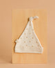 The Organic Baby Hat – Gold Sprig is a soft, white newborn hat featuring tiny yellow and orange floral prints and a knotted top, shown flat on a light wooden board with a neutral beige background.