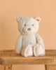 The Steiff Bearzy Teddy Bear Stuffed Plush for Baby, 11 inches, in light beige with gray eyes and nose, made in Germany from ultra soft plush, sits upright on a wooden stool against a peach background.