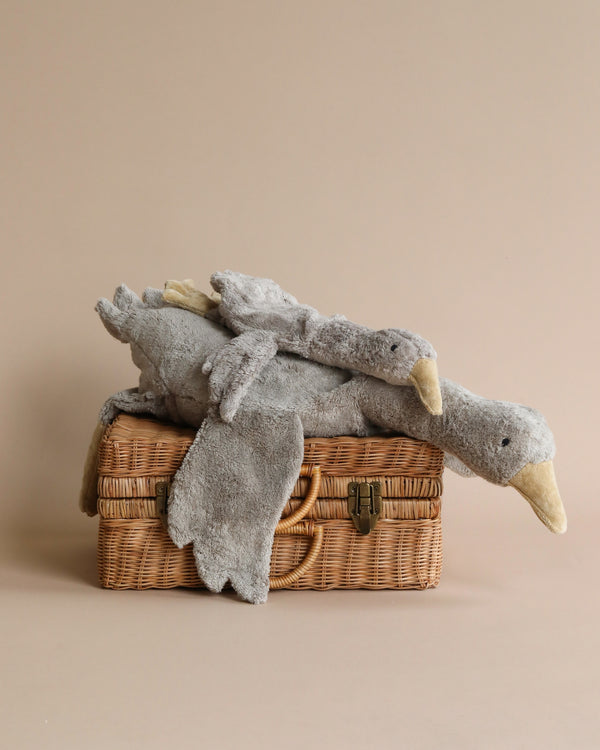 A Senger Naturwelt Cuddly Animal - Grey Goose plush toy resembling a goose, draped over a closed wicker picnic basket, set against a plain beige background.