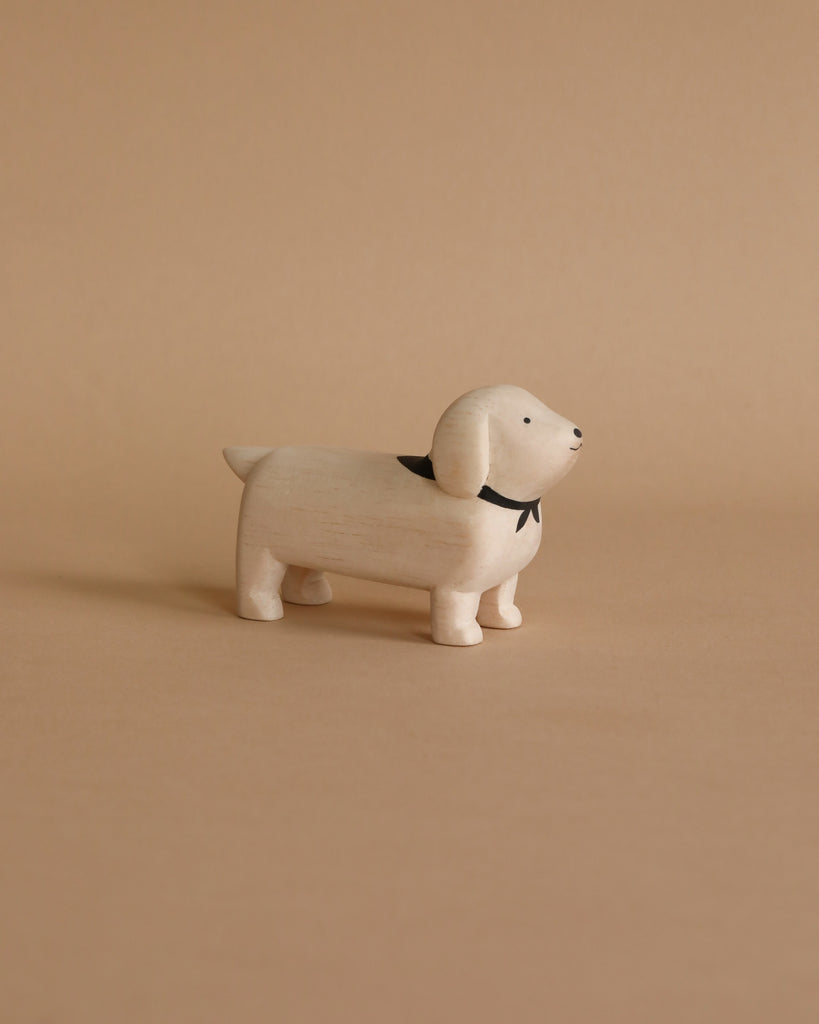 A Handmade Tiny Wooden Animals - White Dachshund Dog with a black collar stands on a neutral beige background. Handcrafted by skilled artisans from Albizia wood, the dog has a simple, minimalist design with a smooth texture and a slight smile on its face.