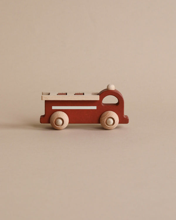 A small, red Mini Wooden Fire Truck toy with white wheels stands against a neutral beige background. The toy is simple in design with clearly visible axles and removable ladder details.