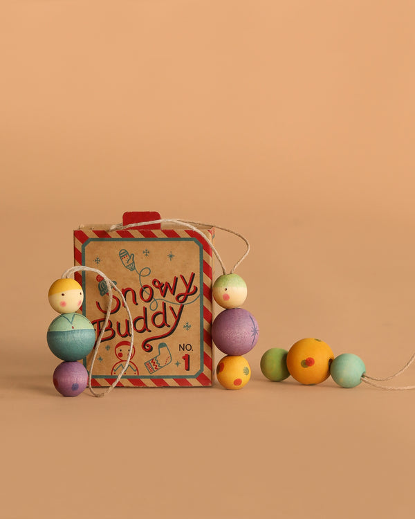 A box labeled "Grapat Ornament Set - Snowy Buddy" sits on a beige background, draped with pastel wooden bead ornaments featuring smiley faces—perfect for adding unique holiday cheer.