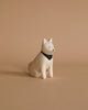 A small, white ceramic dog with a black nose and a painted black bandana around its neck is sitting against a light brown background. The dog's expression is neutral, and its eyes are small black dots, reminiscent of the detail found in Handmade Tiny Wooden Farm Animals - Akita Dog.