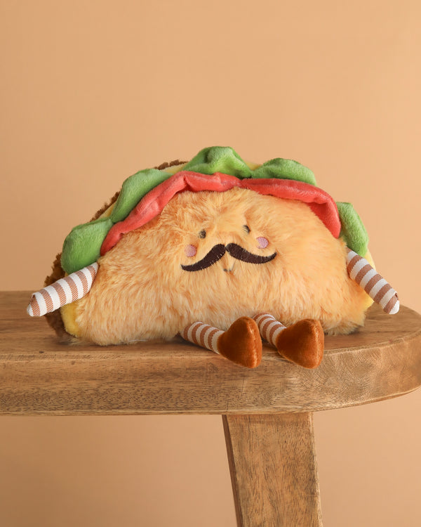 The Paco Taco Plush Toy, shaped like a taco with a mustache, smiling face, and striped arms and legs, sits on a wooden stool against a plain beige background.