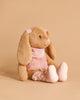 The Steiff Cowgirl Hoppie Rabbit, a plush bunny with floppy ears, light brown fur, and pink accents on its ears, chest, and feet, sits upright against a beige background—a perfect cuddly toy for any collection.