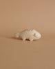 A small, hand-carved Albizia wood handmade tiny wooden animals - chameleon figurine, crafted by skilled artisans in Bali, features simple black line details for eyes, a mouth, and patterned markings on its back. Set against a peach-colored background, the light beige chameleon has a charming curved tail.