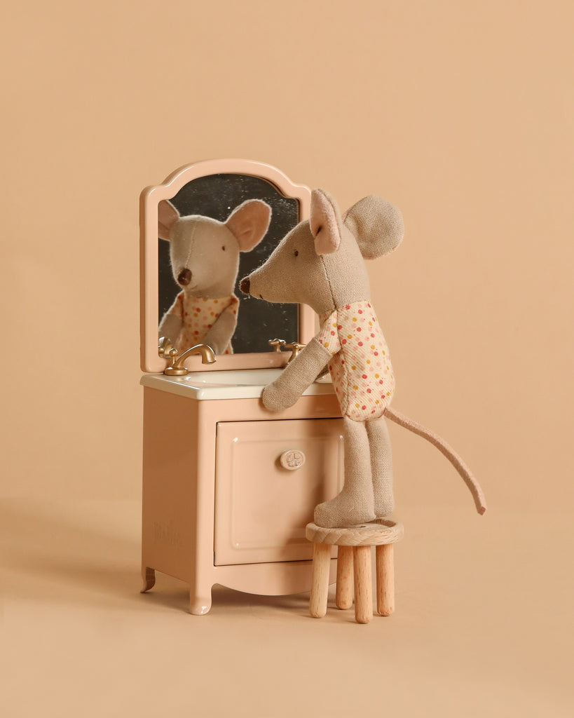The Maileg Little Sister Mouse Vanity Set features a mouse in a floral shirt standing on a stool, gazing into a tiny mirror atop beige furniture—perfect for completing her cozy mouse house against the tan background.