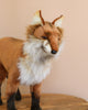 The Standing Red Fox Stuffed Animal is showcased on a wooden surface against a beige background, featuring artisan hand-sewn craftsmanship with reddish-brown, white, and black fur. It has pointed ears and a bushy tail, capturing its unique personality.