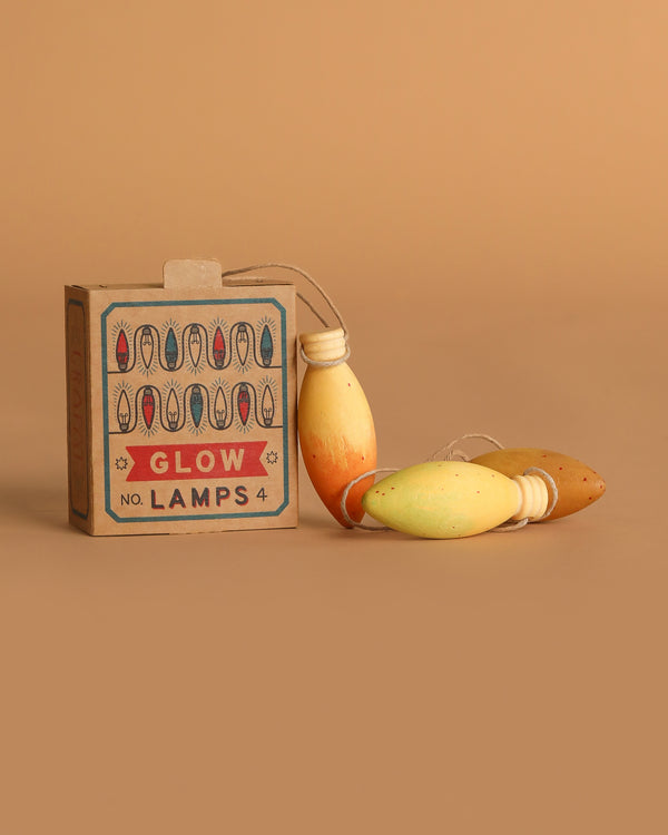 A Grapat Ornament Set - Glow Lamps box sits beside three vibrant, bulb-shaped string lights—unique holiday decorations—on a brown surface with a matching background.