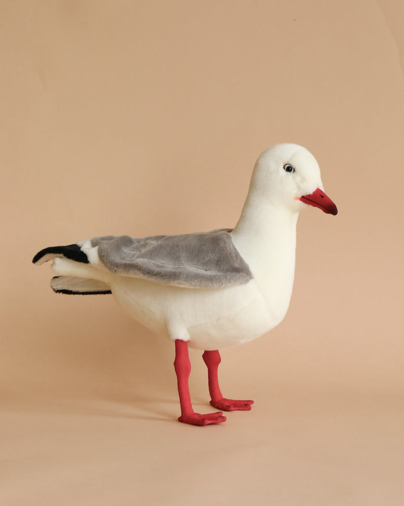 A Seagull Bird Stuffed Animal standing on a plain beige background. The bird, an artisan-crafted toy, features prominent white and gray plumage, a bright red beak, and vivid red legs.