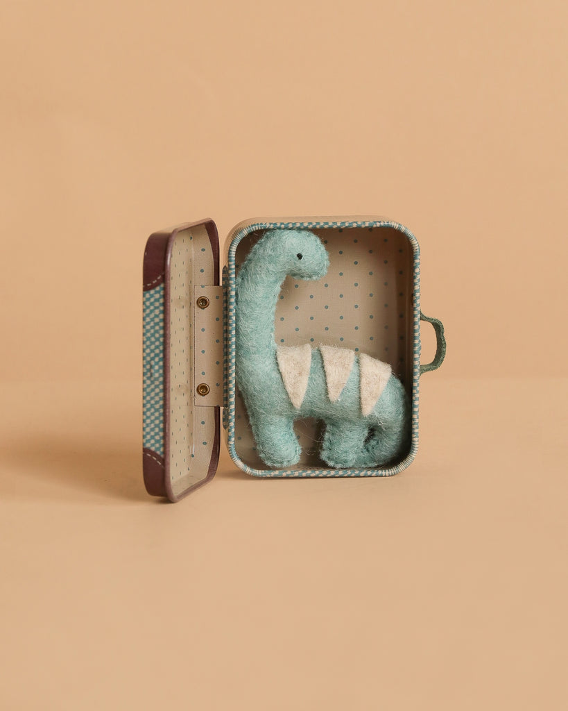 The Tiny Dinosaur Travel Set features a handmade blue felt dinosaur with white details, sitting inside an open Maileg polka dot suitcase against a plain beige background.