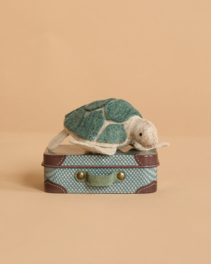 The Tiny Turtle Travel Set features a small, handmade felt turtle with a green shell sitting on a blue patterned Maileg micro suitcase, displayed against a plain beige background.