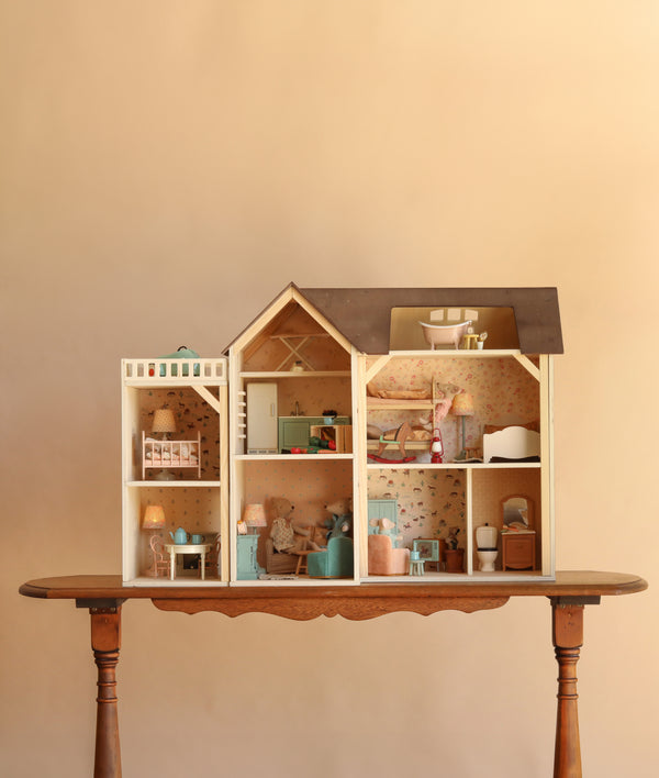 A Maileg Fully Furnished Farmhouse Set + Surprise Gift sits on a wooden table, featuring multiple rooms decorated with miniature furniture and wallpaper—including charming dollhouse accessories and Maileg Mouse Furniture—with a cozy kitchen, bathroom, bedroom, and inviting living spaces.