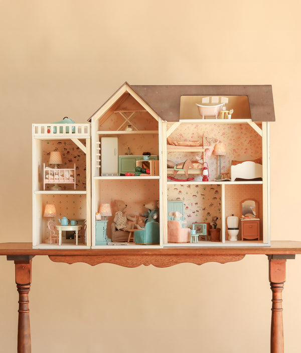 The Maileg Fully Furnished Farmhouse Set + Surprise Gift is displayed on a wooden table against a plain beige background. This detailed dollhouse features multiple rooms furnished with charming Maileg accessories, including tiny pastel-colored furniture, lamps, and wallpaper.