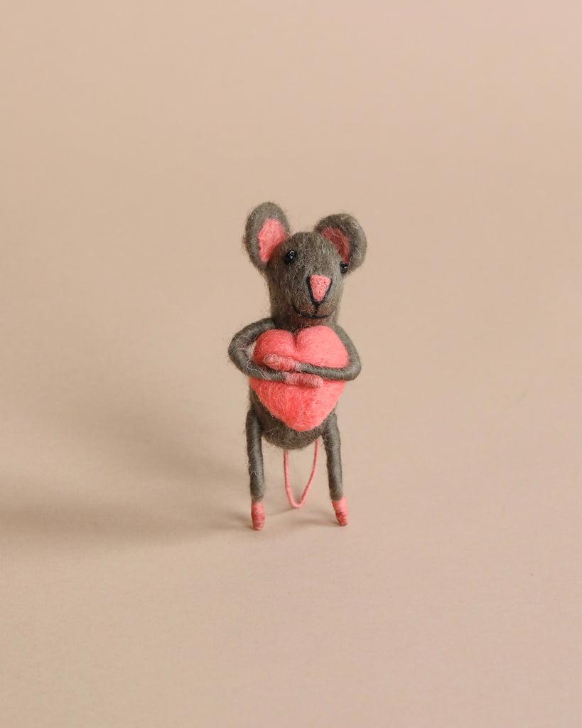A small hand-felted grey mouse holding a pink heart in its arms. Photographed on a beige background.