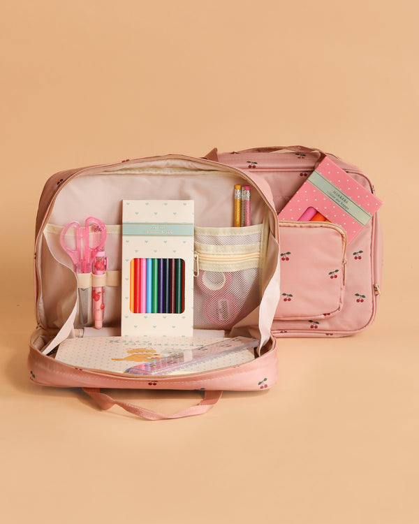 The Konges Sloejd Holiday Essentials Bag - Cherry, an open pink kids travel bag with cherry patterns, showcases stationery items against a peach background. A matching backpack appears in the background—ideal for holiday essentials.