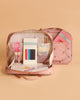 The Konges Sloejd Holiday Essentials Bag - Cherry, an open pink kids travel bag with cherry patterns, showcases stationery items against a peach background. A matching backpack appears in the background—ideal for holiday essentials.