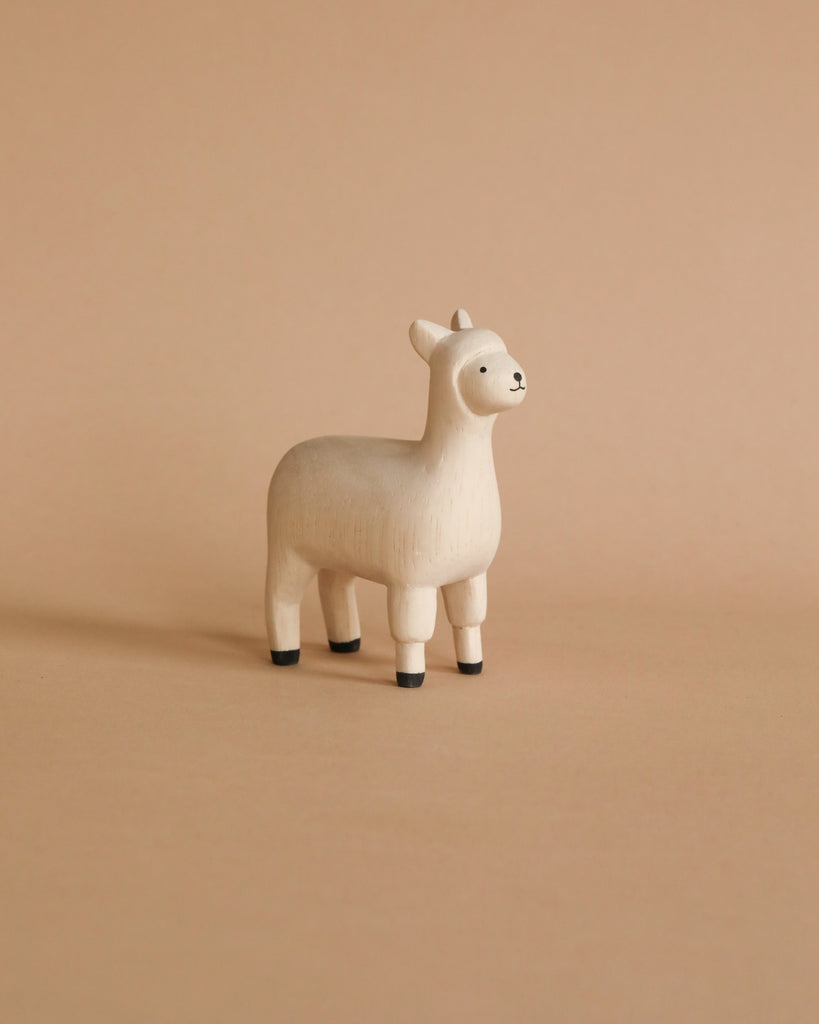 A small, white wooden figurine of a llama, **Handmade Tiny Wooden Farm Animals - Alpaca**, hand carved from Albizia wood by artisans in Bali, stands against a plain, beige background. The figurine has black-tipped feet and a simple, cute expression on its face.