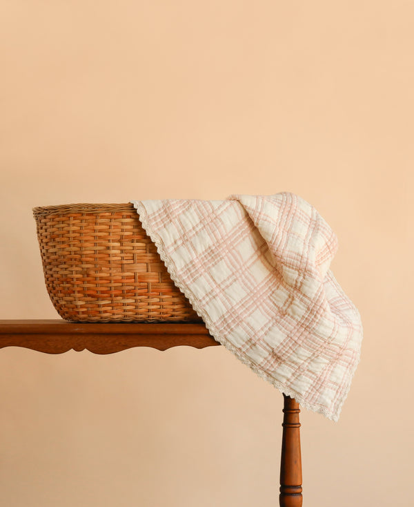 A wicker basket with a pastel plaid cloth draped over its edge sits on a wooden table against a peach-colored background, complemented by the cozy Organic Cotton Baby Blanket - French Plaid, Petal, made in Portugal.