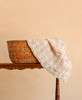 A wicker basket with a pastel plaid cloth draped over its edge sits on a wooden table against a peach-colored background, complemented by the cozy Organic Cotton Baby Blanket - French Plaid, Petal, made in Portugal.