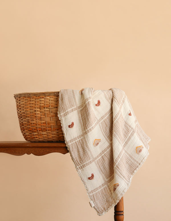 An Organic Cotton Baby Blanket - Farm, adorned with small animal designs and a delicate crochet trim, is draped over the edge of a woven basket resting on a wooden table, set against a plain tan background.