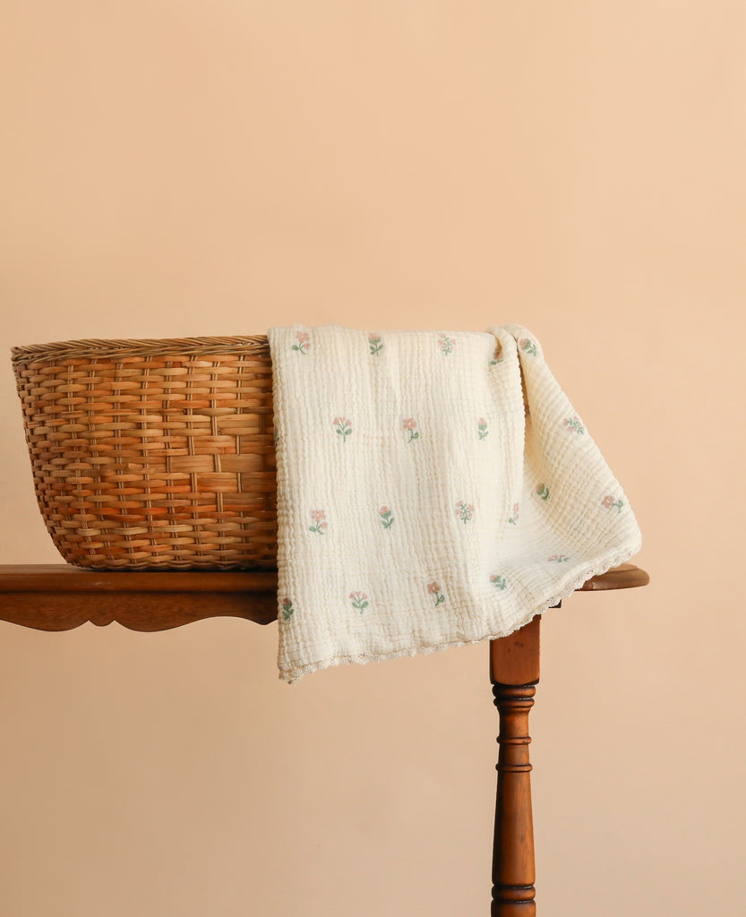 The Organic Cotton Layered Gauze Blanket - Dahlia, featuring a cream-colored floral pattern, is draped over the side of a woven basket resting on a wooden table. The background is a solid light peach color.