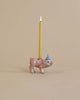 A Pig Cake Topper shaped like a pink pig wearing a blue and white striped party hat with a lit yellow candle on its back, set against a plain beige background. This porcelain keepsake is handcrafted.
