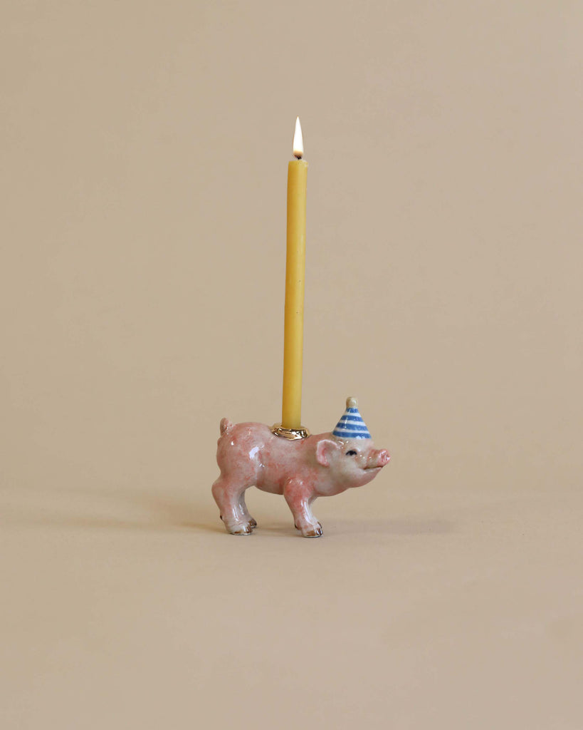 A Pig Cake Topper shaped like a pink pig wearing a blue and white striped party hat with a lit yellow candle on its back, set against a plain beige background. This porcelain keepsake is handcrafted.