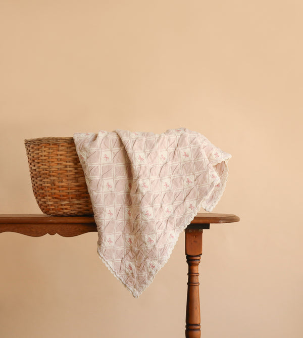 An Organic Cotton Baby Blanket - Pink Bows is draped gracefully over the edge of a wicker basket sitting on a wooden table, its delicate pink bow patterns and soft hues standing out against a plain beige background.