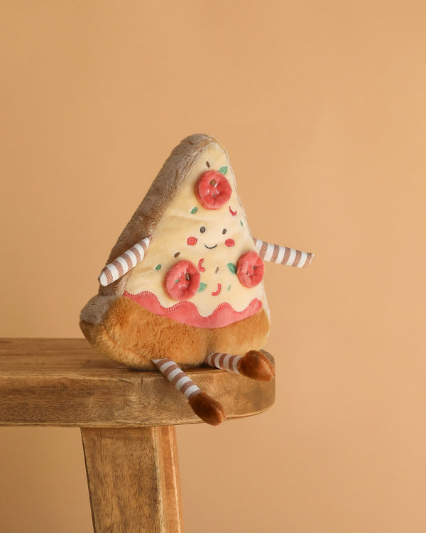 The Cosimo Pizza Slice Plush Toy, featuring a smiling face, felt tomato and sprinkle toppings, plus striped arms and legs, sits on a wooden stool against a plain beige background—an adorable gift for all ages.