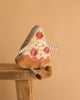 The Cosimo Pizza Slice Plush Toy, featuring a smiling face, felt tomato and sprinkle toppings, plus striped arms and legs, sits on a wooden stool against a plain beige background—an adorable gift for all ages.
