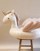 An Inflatable Unicorn Float in beige and white, with handles on the neck, placed upright on a wooden stool against a neutral beige backdrop.