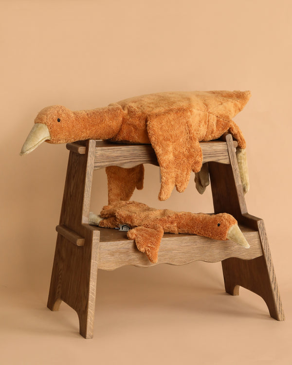 Two large Senger Naturwelt Cuddly Animal - Rust Geese are draped over a two-level wooden step stool against a beige background. The plush geese look soft and relaxed, with their heads and limbs hanging down.