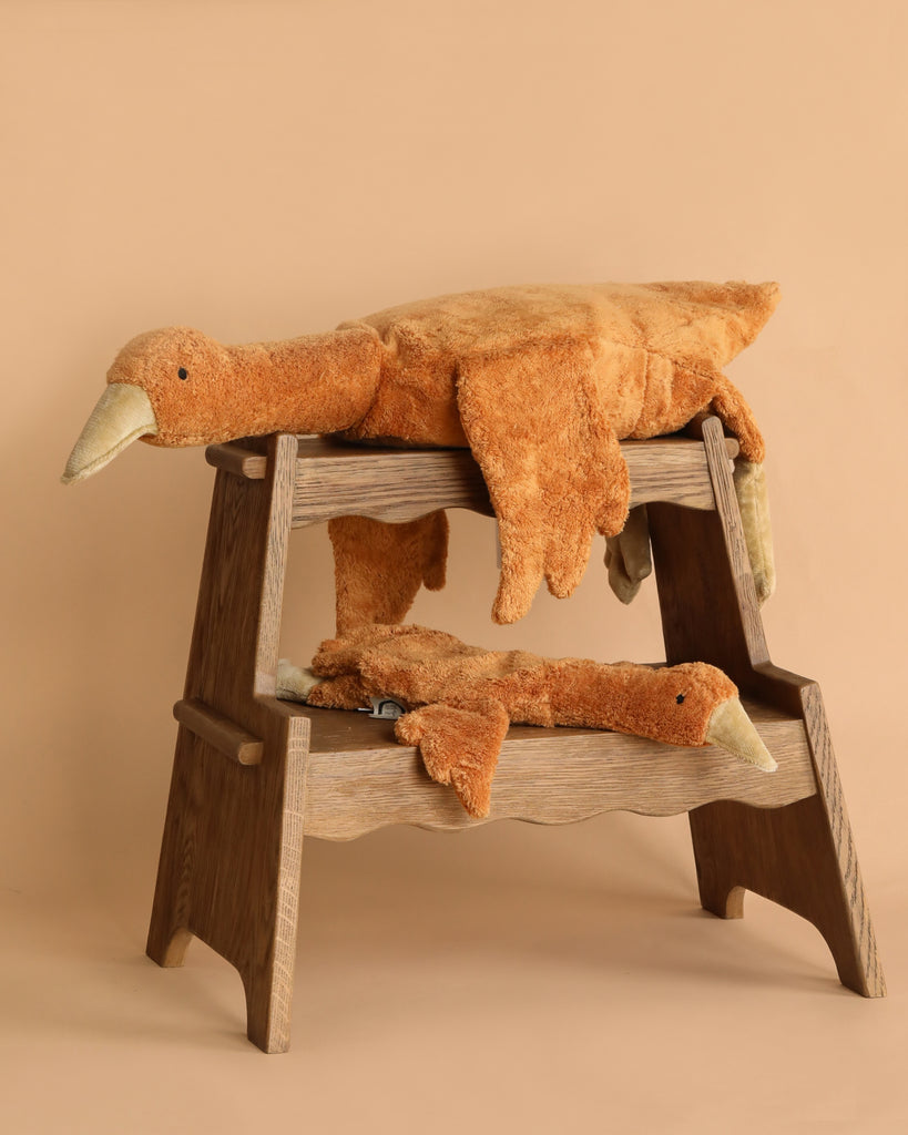 Two large Senger Naturwelt Cuddly Animal - Rust Geese are draped over a two-level wooden step stool against a beige background. The plush geese look soft and relaxed, with their heads and limbs hanging down.