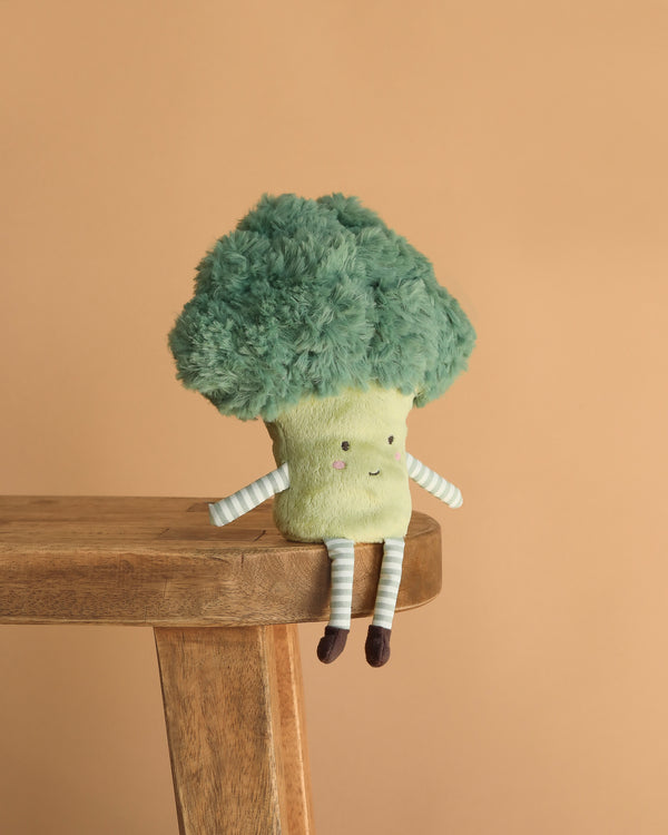The Bro Broccoli Plush Toy, featuring a smiling face and striped arms and legs, sits on a wooden bench against a beige background—an adorable stuffed vegetable and perfect cuddle buddy.