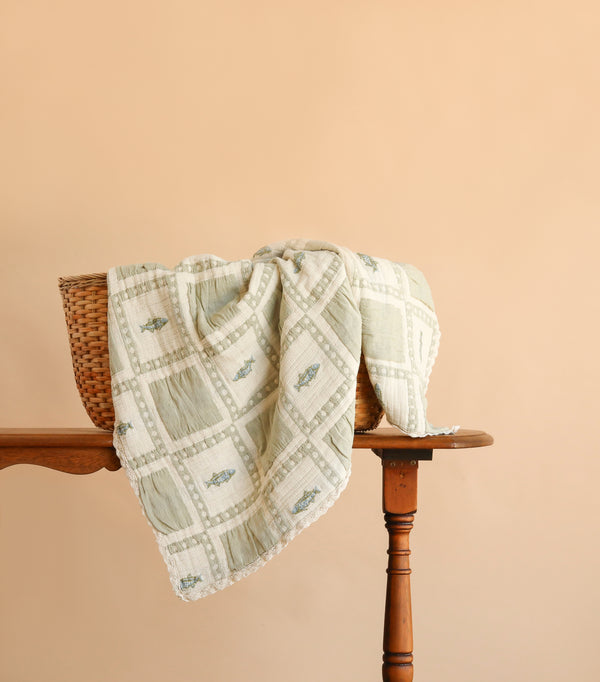An Organic Cotton Baby Blanket - Fishing is draped over a wicker basket that sits atop a wooden table against a plain peach background. The light-colored, patterned blanket—crafted in Portugal—introduces a touch of gentle elegance to the scene.