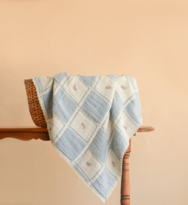 The Organic Cotton Baby Blanket - Spring Bloom, featuring blue and white checks adorned with small pink flowers, is draped over a wicker basket on a wooden table against a beige background.