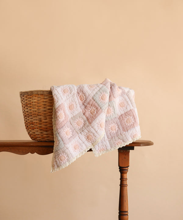A woven basket sits on a wooden table with the Organic Cotton Quilt - Sundrop, hand dyed in pastel pink and white from organic cotton, draped over the edge; the background is a soft beige color.