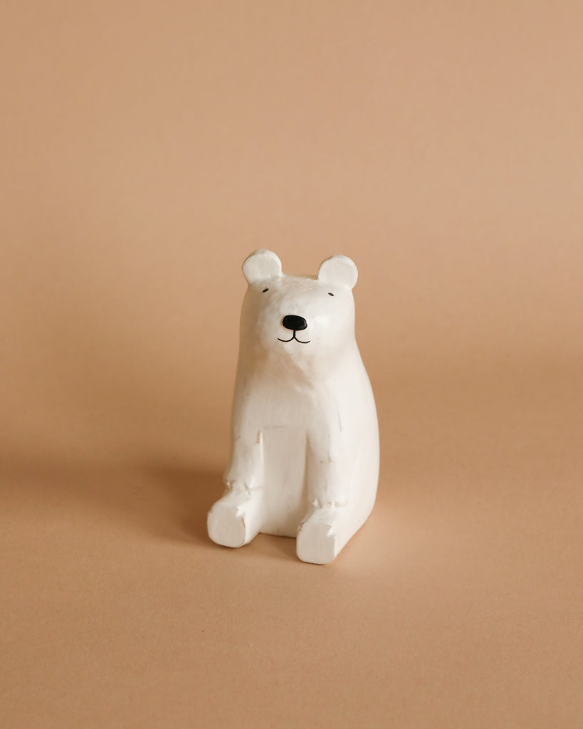 A small, white ceramic bear figurine sits upright on a light brown surface against a neutral background. The bear has minimalistic, black facial features and a simplistic design, giving it a cute and modern aesthetic. Its charm complements Handmade Tiny Wooden Polar Bears beautifully.