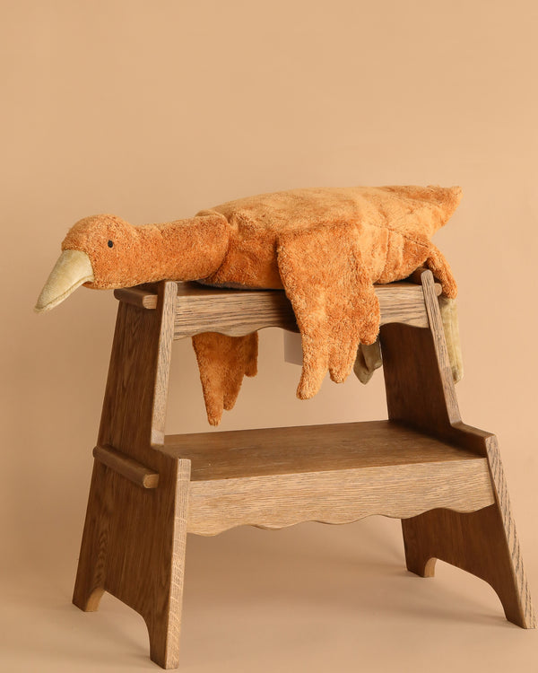 The Senger Naturwelt Cuddly Animal - Rust Goose, a large plush toy with an orange-brown body and beige beak, lies draped over a wooden step stool against a beige background.