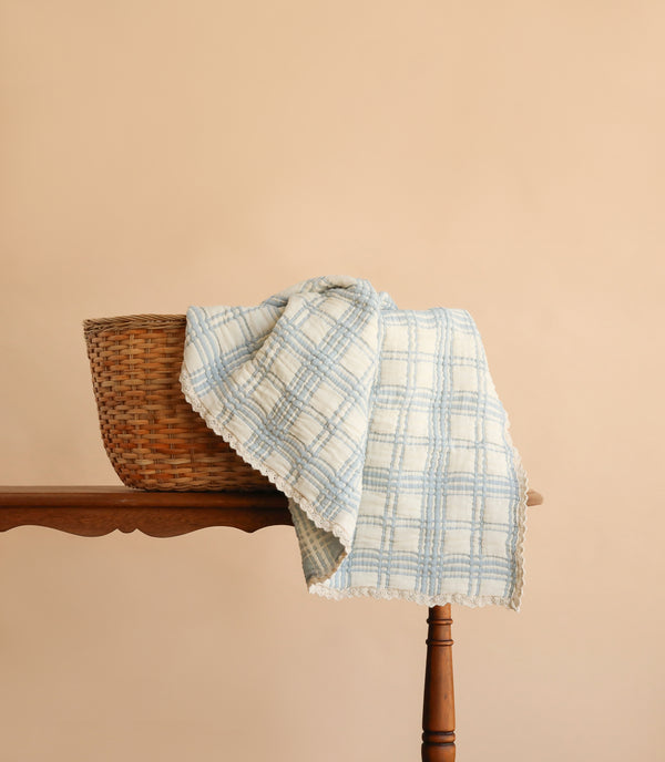A woven basket rests on a wooden table with the Organic Cotton Baby Blanket - French Plaid, Powder Blue draped over it, set against a plain beige background.
