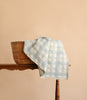 A woven basket rests on a wooden table with the Organic Cotton Baby Blanket - French Plaid, Powder Blue draped over it, set against a plain beige background.