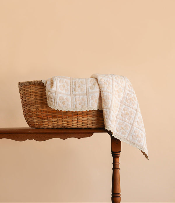 The Organic Cotton Quilt - Daffodil, featuring a cream-colored floral pattern and hand-dyed from organic cotton, is draped over the side of a woven basket on a wooden table against a plain beige background.